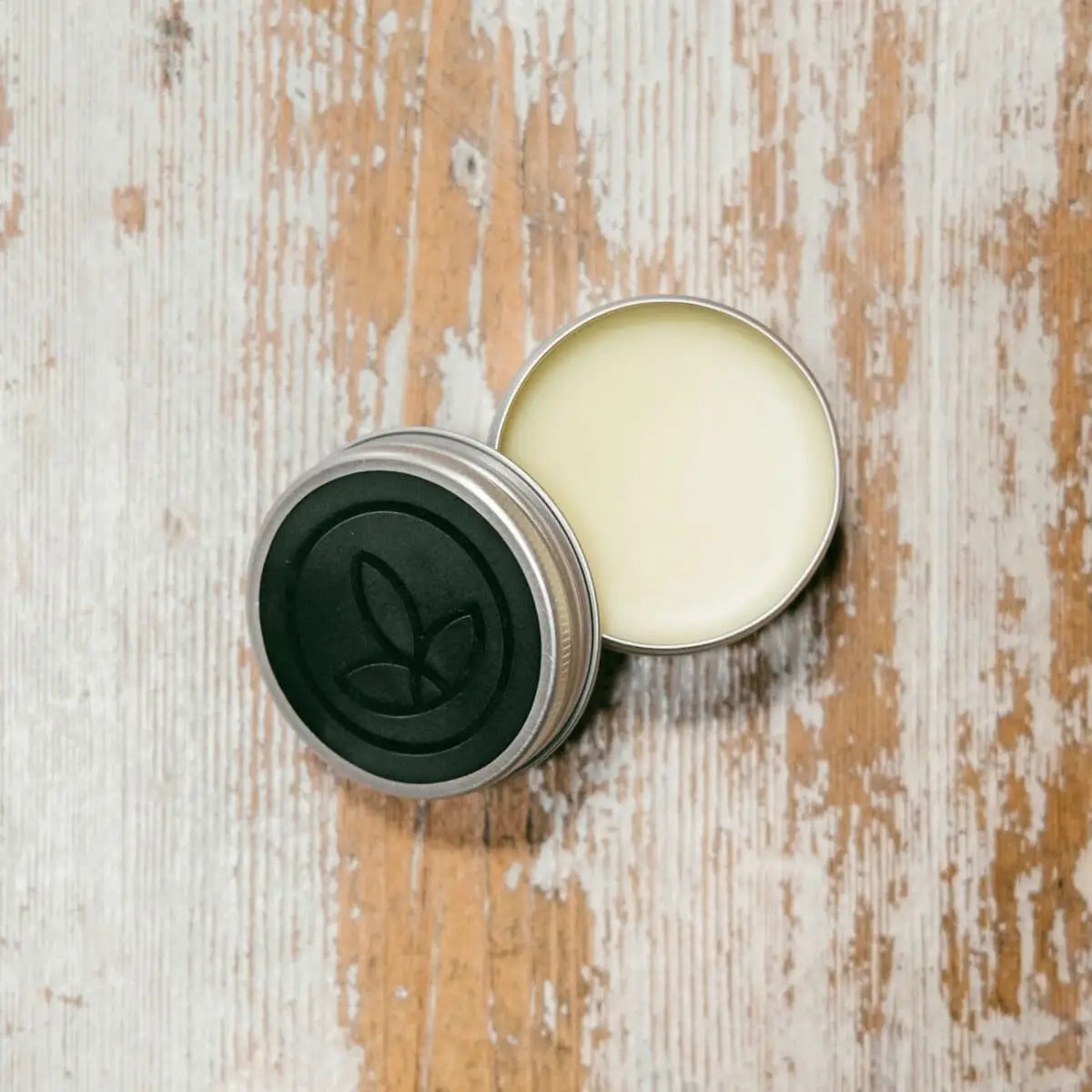 A small, round metal tin of 1000MG CBD Balm with a black lid beside it reveals a solid balm inside, placed on a rustic, weathered wooden surface.