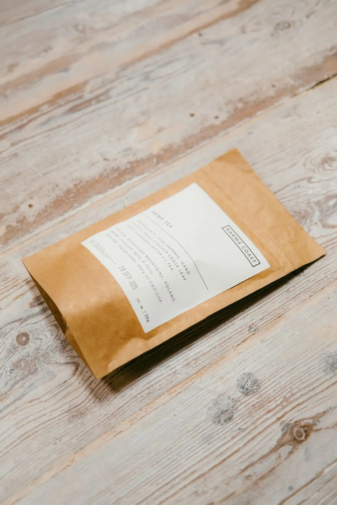 KARMA ORGANIC CBD TEA 2 A brown paper pouch with a white label lies on a light wooden surface. The label features printed text, which is not clearly legible in the image, suggesting this could be packaging for KARMA ORGANIC CBD TEA.