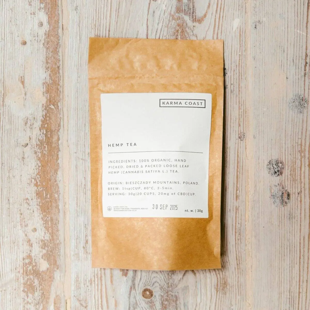 A brown paper packet of KARMA ORGANIC CBD TEA rests on a light wooden surface. The white label displays the ingredients and expiry date. Ideal for those seeking CBD tea UK options, the sealed packet is centrally positioned.