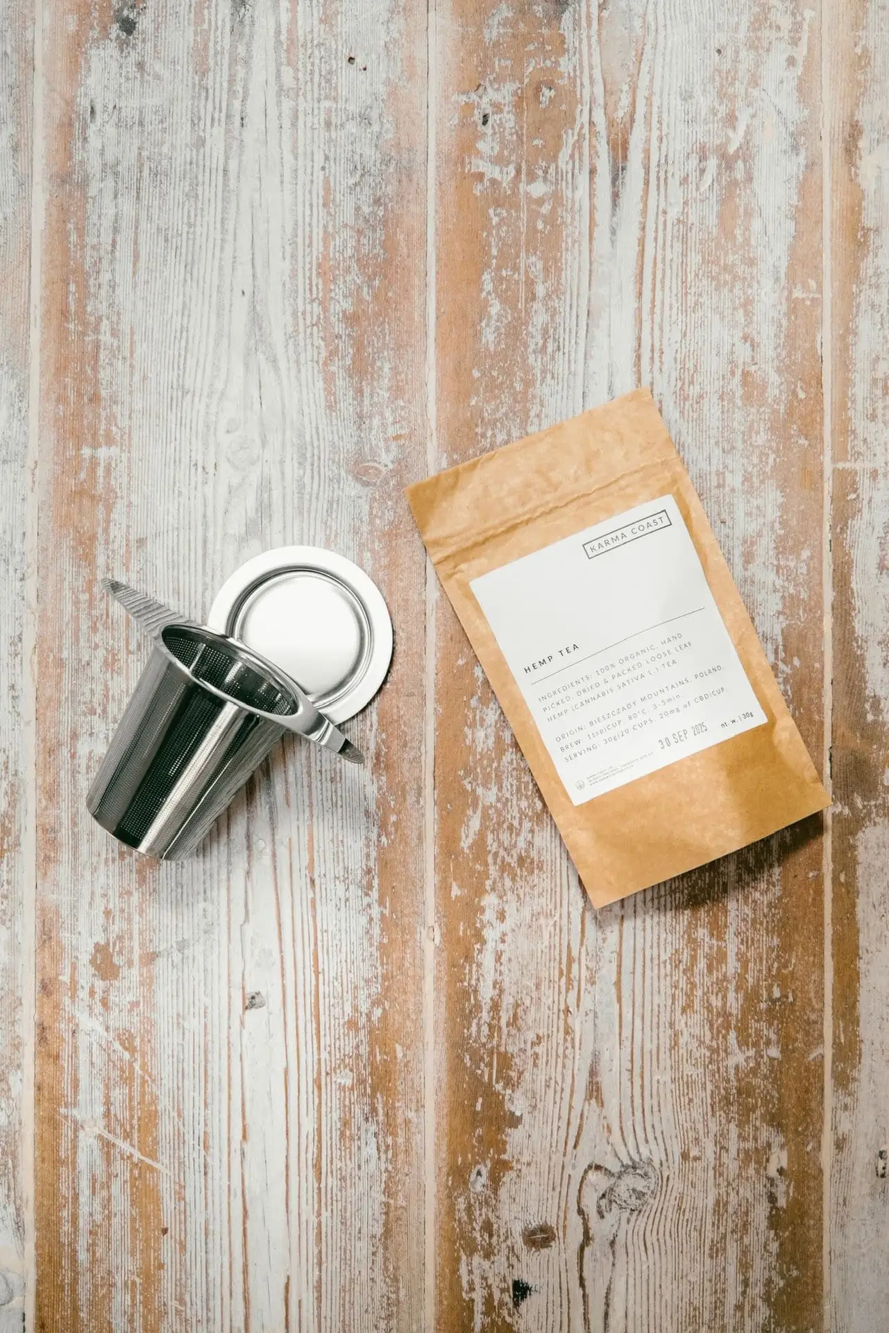 KARMA ORGANIC CBD TEA 3 A metal tea infuser with a lid sits next to a brown paper bag of KARMA ORGANIC CBD TEA on a rustic wooden surface, evoking the calming ritual of preparing CBD tea UK for a soothing evening unwind.