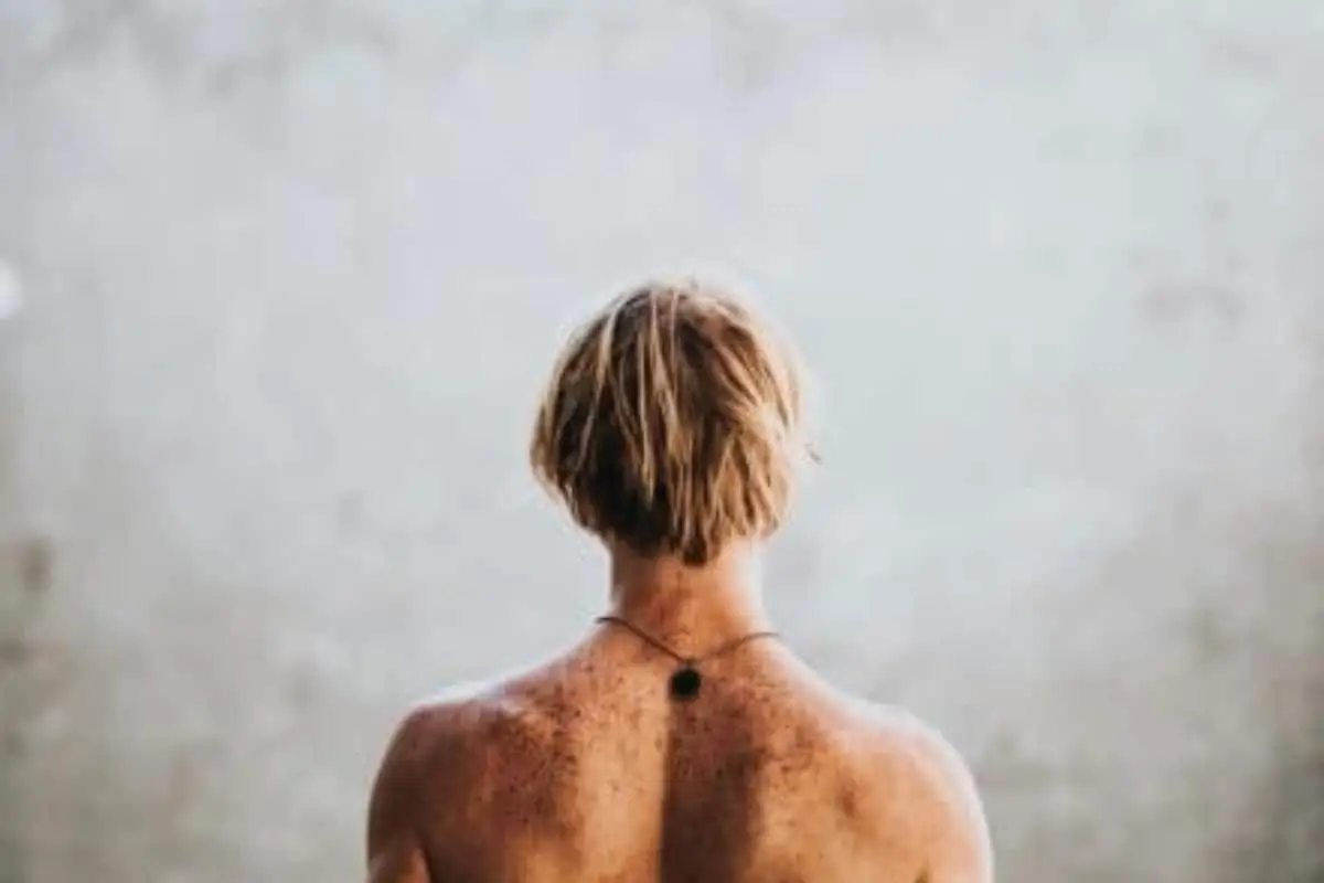 A person with short, light brown hair is seen from behind, showing their bare freckled back and a necklace with a small pendant against a plain, light background—a serene image that subtly hints at themes of CBD and inflammation.