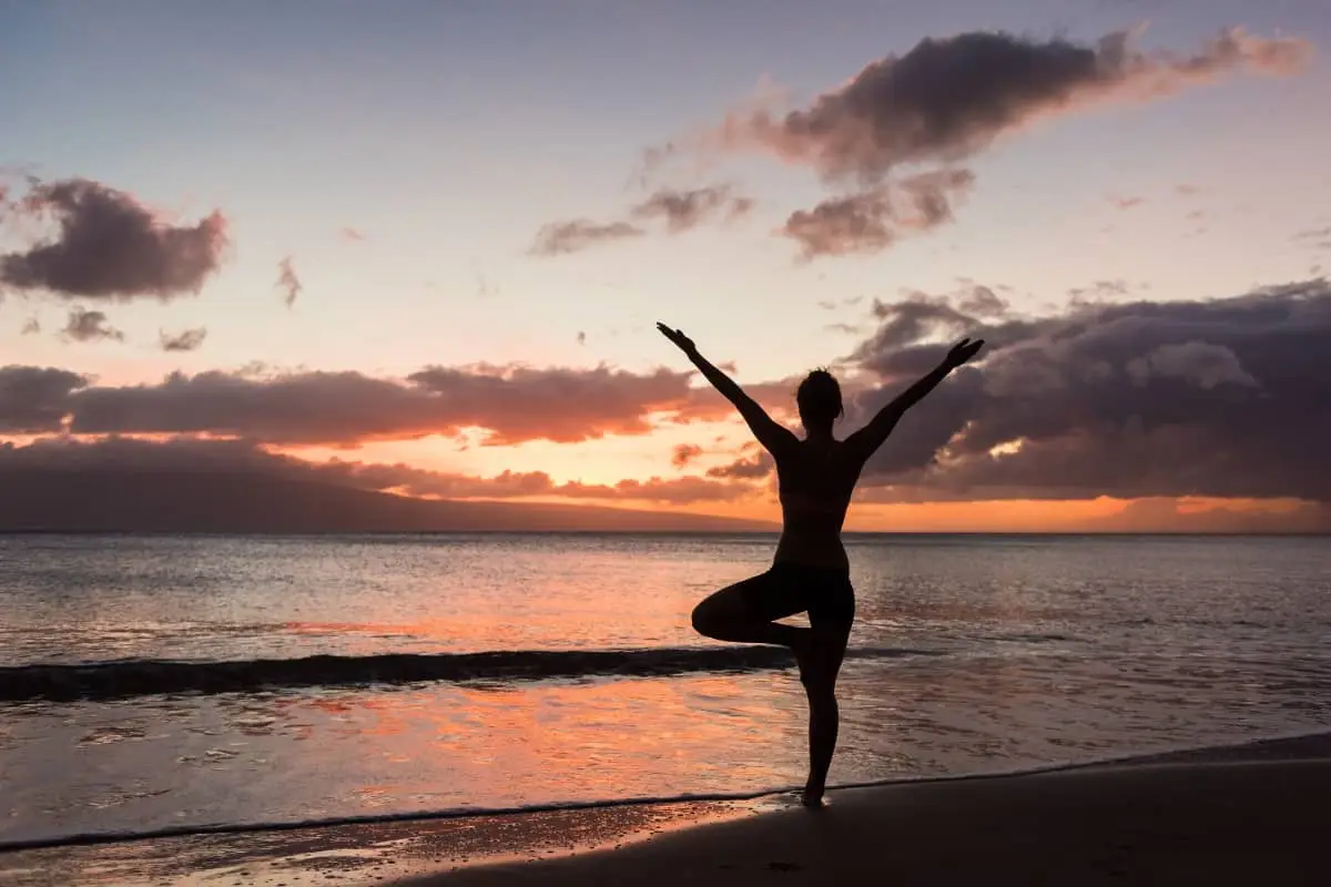 A person stands in a yoga pose on the beach at sunset, silhouetted against a colourful sky with clouds and calm ocean waves in the background, embracing the serenity of nature and the benefits of CBD for wellbeing.