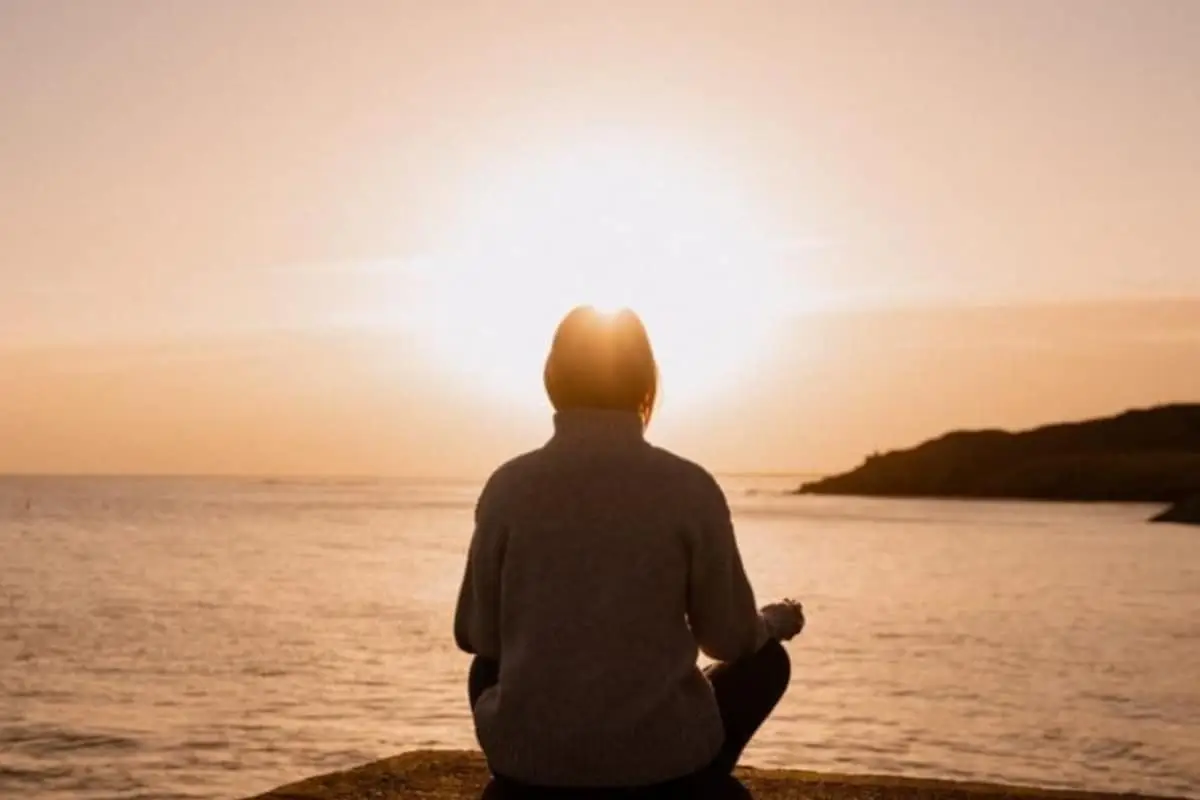 A person sits cross-legged on a rock by the water, facing a bright sunset. The sky is orange, and the silhouette of distant hills is visible. The scene feels calm and peaceful, offering a quiet escape from the effects of stress.