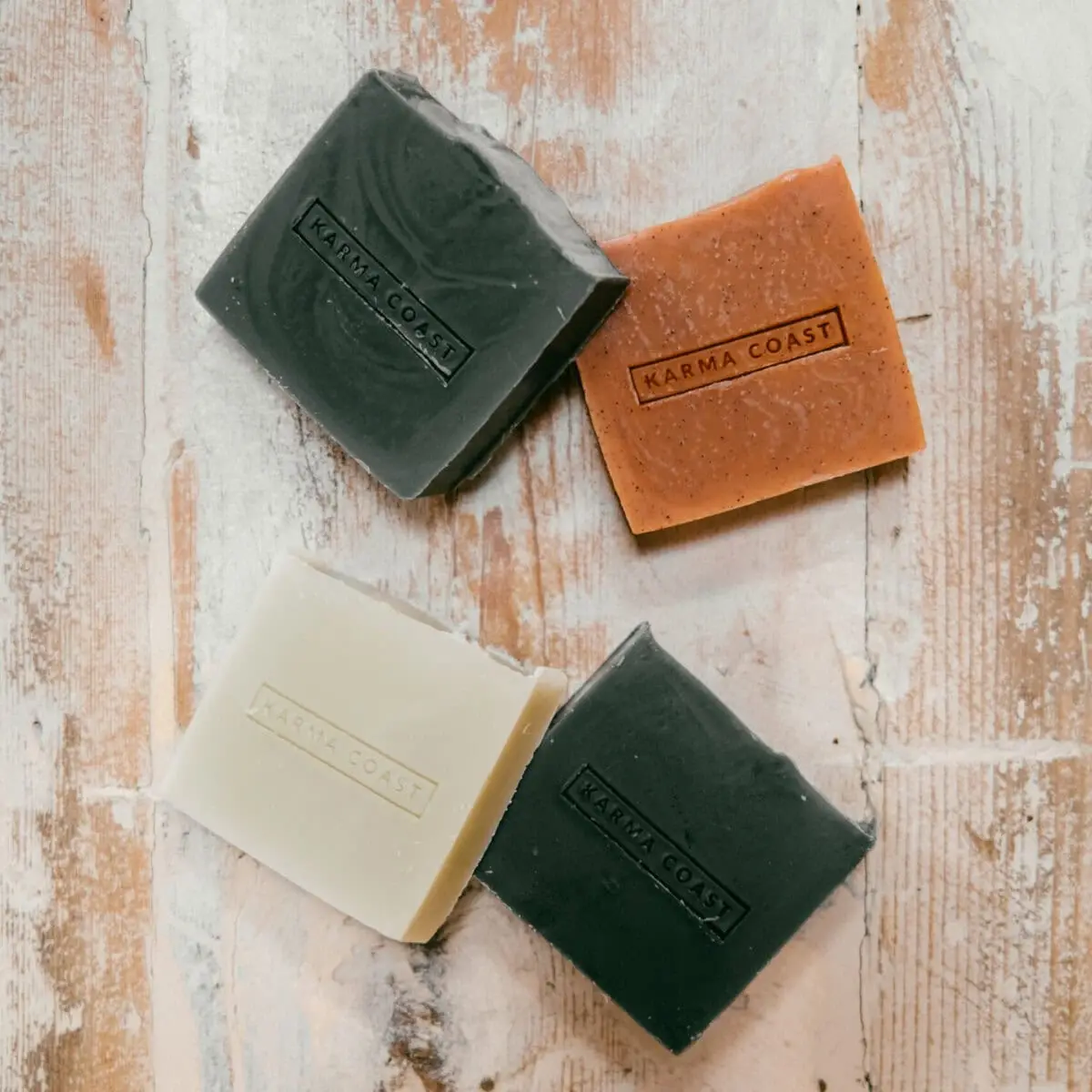 Four rectangular bars of Karma Hemp Soap—two dark green, one white, and one orange—are arranged in a loose diamond shape on a rustic wooden surface. Each bar features an embedded Karma Coast label.