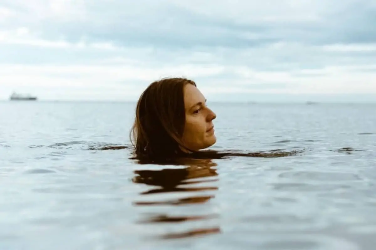 A person with long brown hair is partially submerged in calm water, looking to the left. The sky is cloudy, and a ship is visible in the distant background.