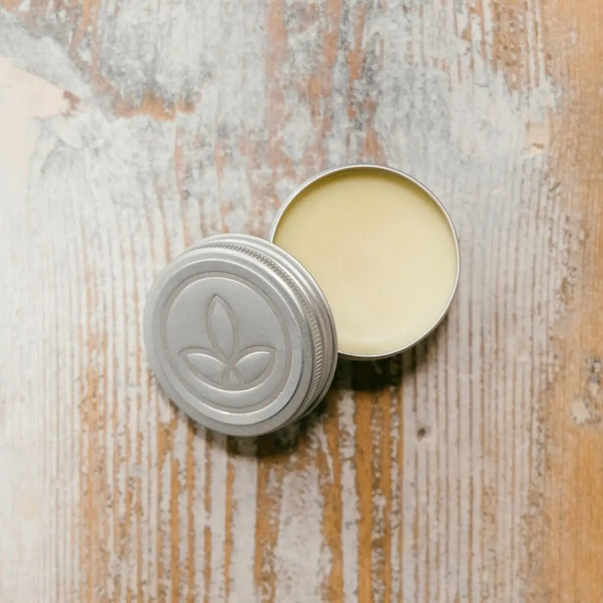 A small, round tin labelled "250MG CBD Balm" with a lid featuring a simple leaf design, containing a pale yellow balm, sits on a rustic, light-coloured wooden surface.