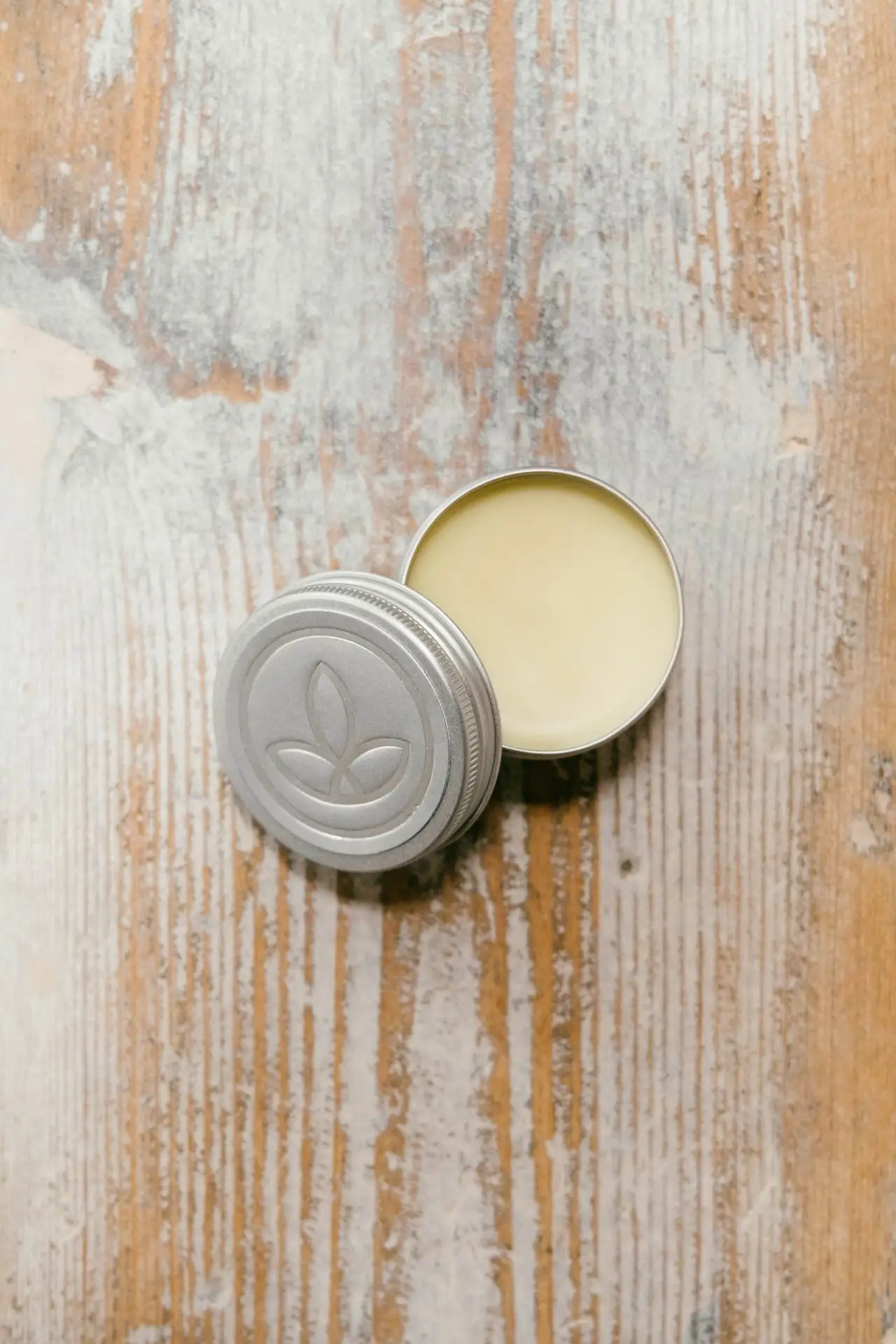 250MG CBD Balm 1 A small, round tin labelled "250MG CBD Balm" with a lid featuring a simple leaf design, containing a pale yellow balm, sits on a rustic, light-coloured wooden surface.