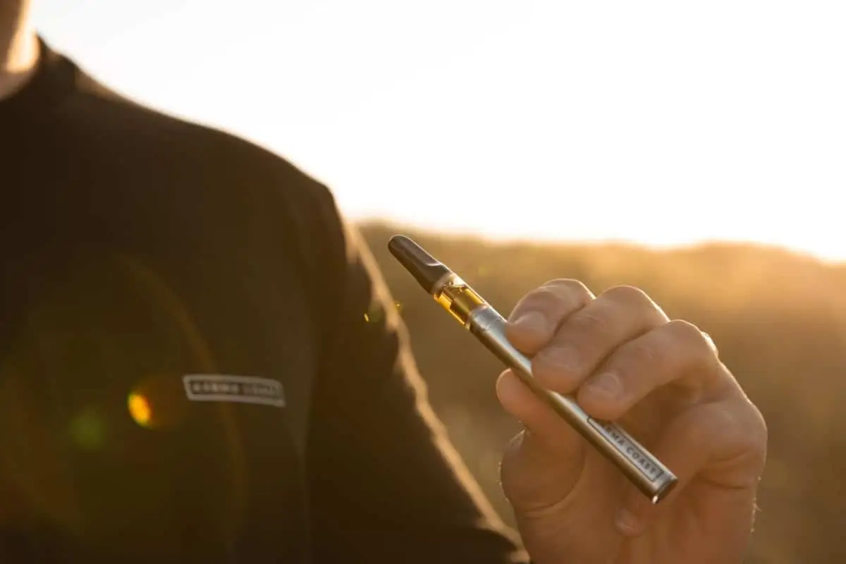 A person holds a vape pen outdoors at sunset, with warm sunlight in the background. Only the hand and part of the torso are visible.