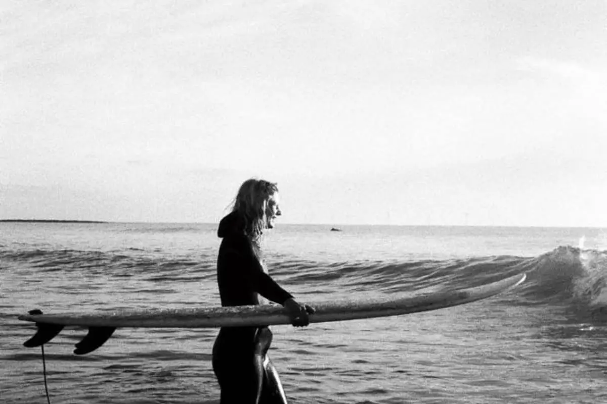 A person in a wetsuit stands in shallow sea water holding a long surfboard, facing small waves. The scene is in black and white.