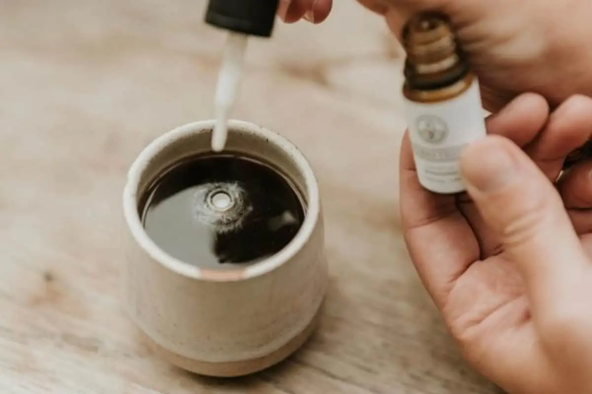 A person holds a small bottle—possibly containing CBD and alcohol—with a dropper poised over a ceramic cup filled with a dark liquid, perhaps coffee or tea, about to add drops from the bottle.