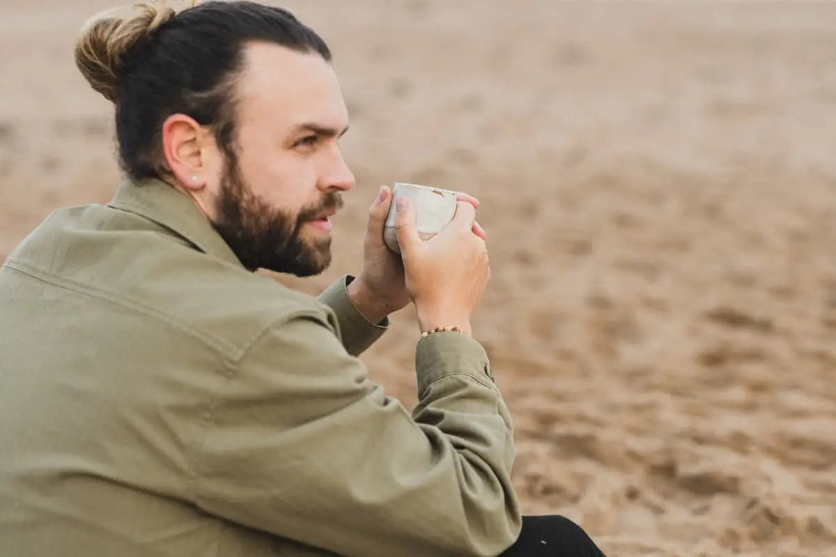 A man with a beard and hair in a bun sits on the sand, holding a mug with both hands and looking thoughtfully into the distance, embodying a quiet moment of reflection and mental health awareness.