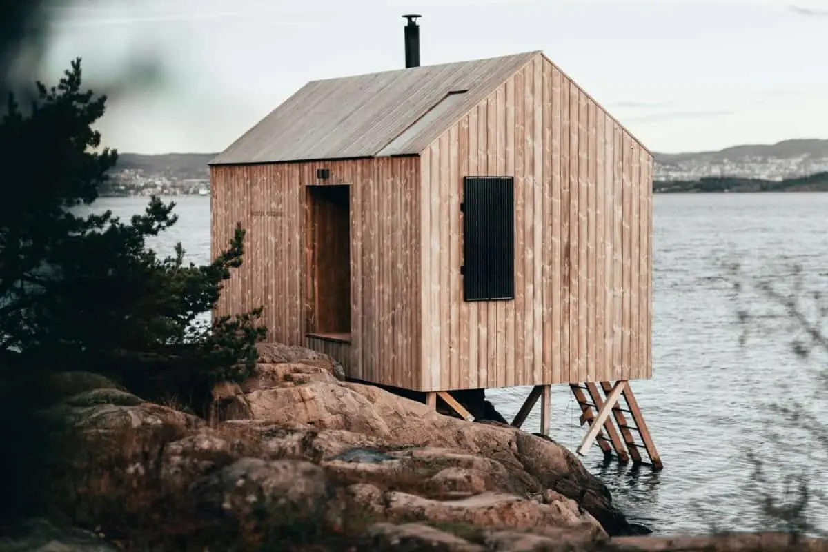 A small wooden cabin with a slanted roof stands on stilts over rocky ground by the water’s edge, surrounded by trees, with distant buildings visible across the water—an ideal retreat for enjoying the benefit of sauna after a day spent in nature.