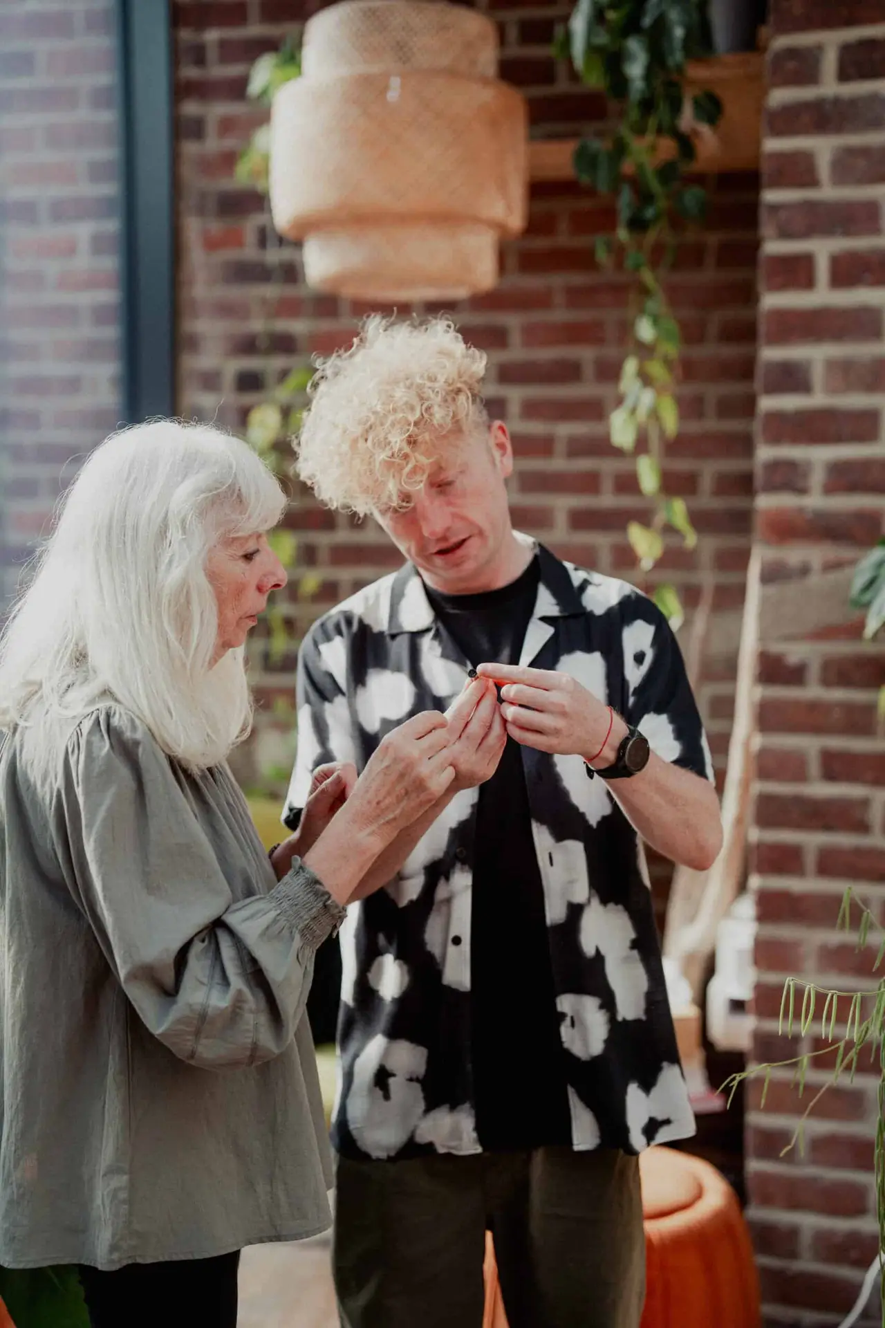 An older woman and a younger man stand indoors by brick walls and greenery, examining a small object together. The woman has long white hair; the man has curly blond hair and wears a black-and-white patterned shirt.