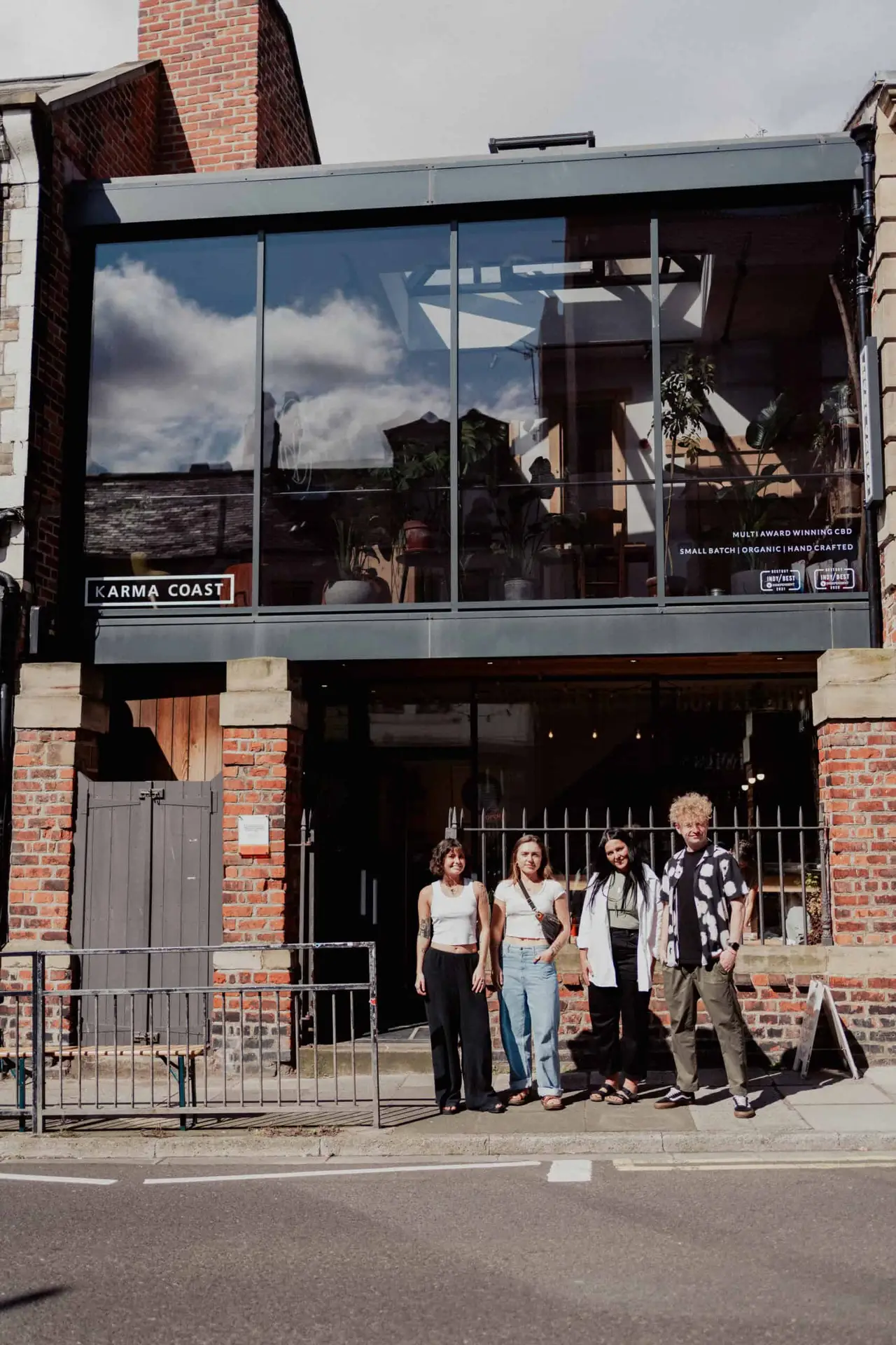 Four people stand in front of a modern brick building with large glass windows and a sign reading “Karma Coast.” The sky’s reflection is visible in the windows above them.