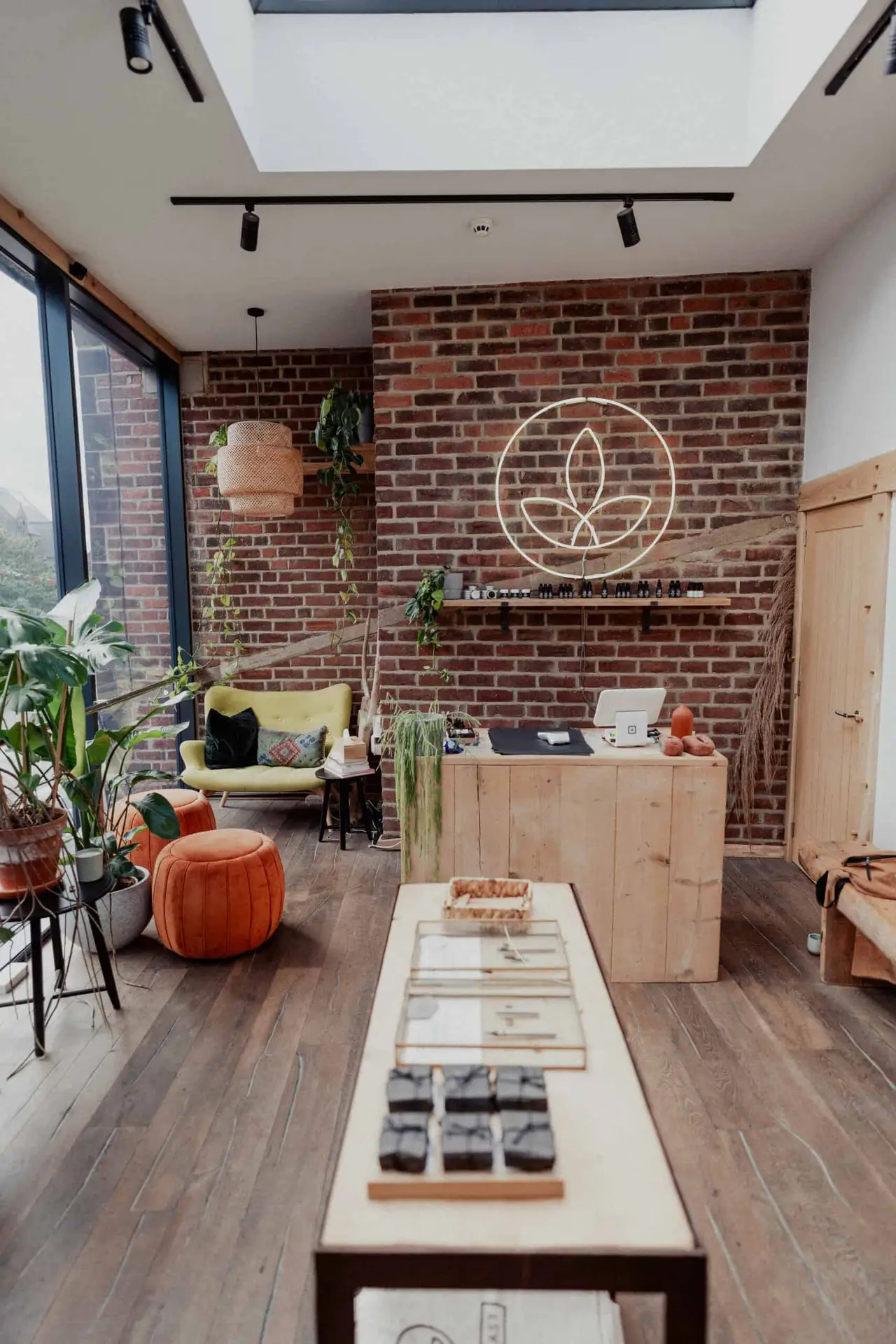 A cosy, modern shop with brick walls, indoor plants, wooden furniture, a skylight, and a neon leaf sign. A counter, lounge chair, orange pouffe, and display table with products are visible.
