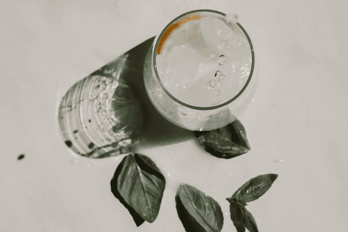 Finding Balance: The Benefits of Reducing Alcohol and How CBD Can Help 1 A glass of clear liquid with ice and a citrus slice, casting a shadow, sits on a light surface near fresh green leaves, possibly basil—an inviting reminder of the benefits of reducing alcohol in your daily routine.