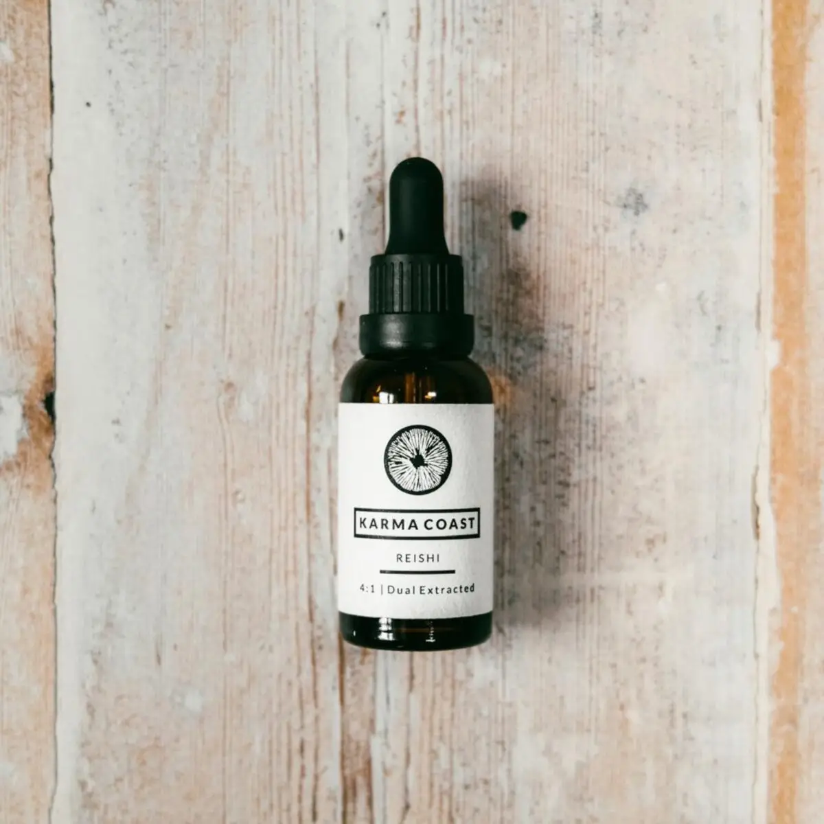 A brown glass dropper bottle labelled Reishi Dual Extract Tincture rests on a light, rustic wooden surface. The label features a circular logo and text describing the product as dual extracted.
