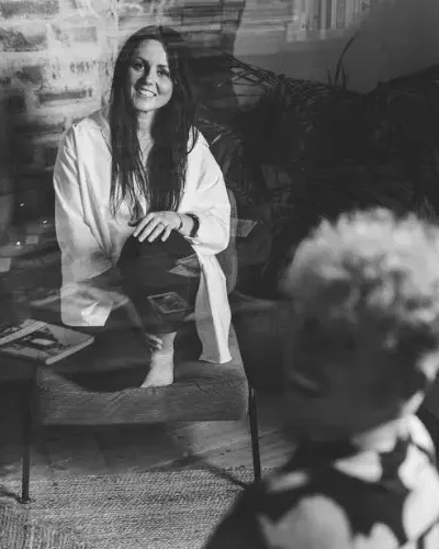 Black and white photo of a woman with long hair smiling and sitting on a chair, with another person blurred in the foreground; a plant and a magazine are visible nearby.